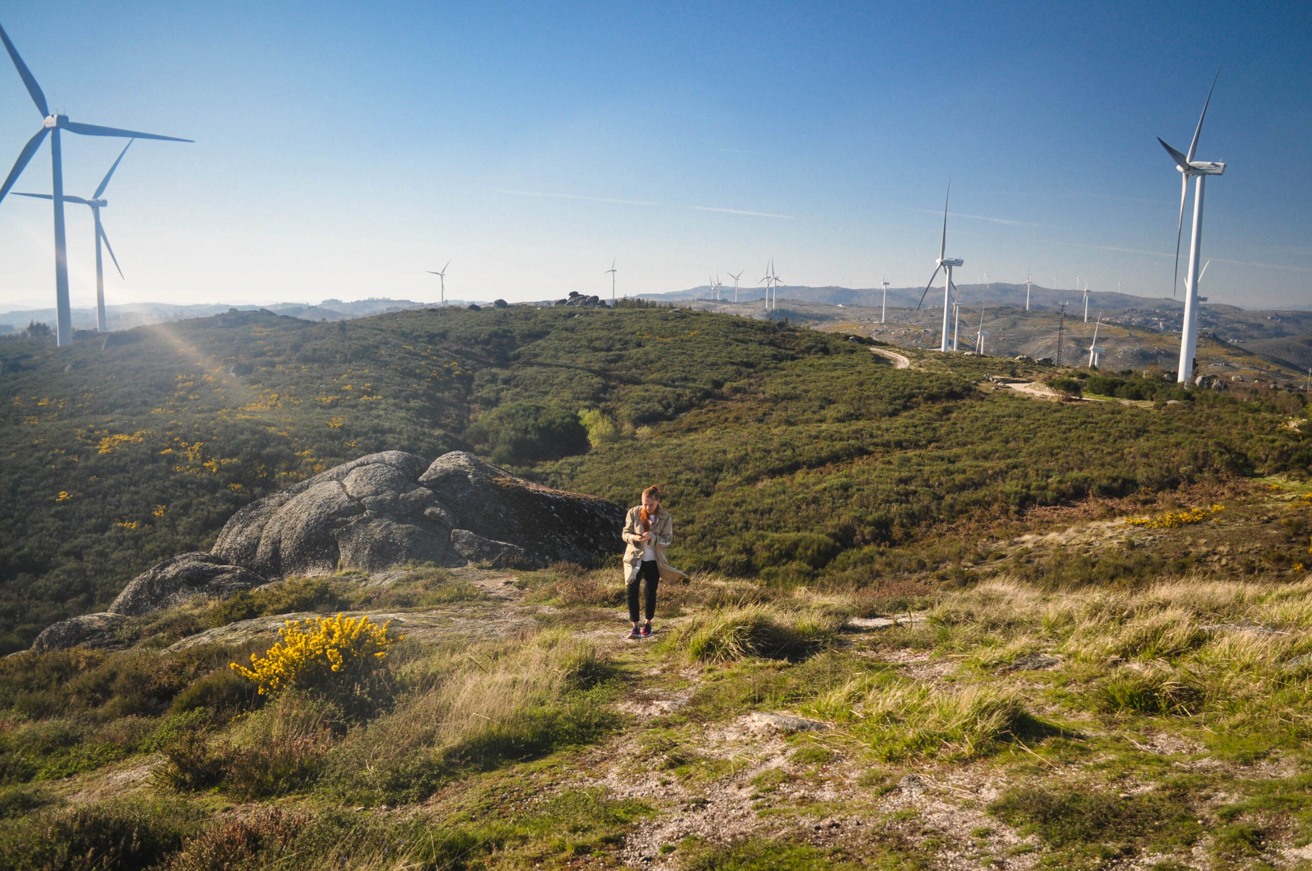 The girl in the beige coat goes on a green hill behind electric wind turbine. The Flintstone House Portugal